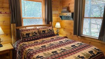 Un dormitorio de cabaña de madera con una cama adornada con colcha temática de osos, dos ventanas con persianas, una mesita de noche con lámpara y un estante montado en la pared con un letrero.