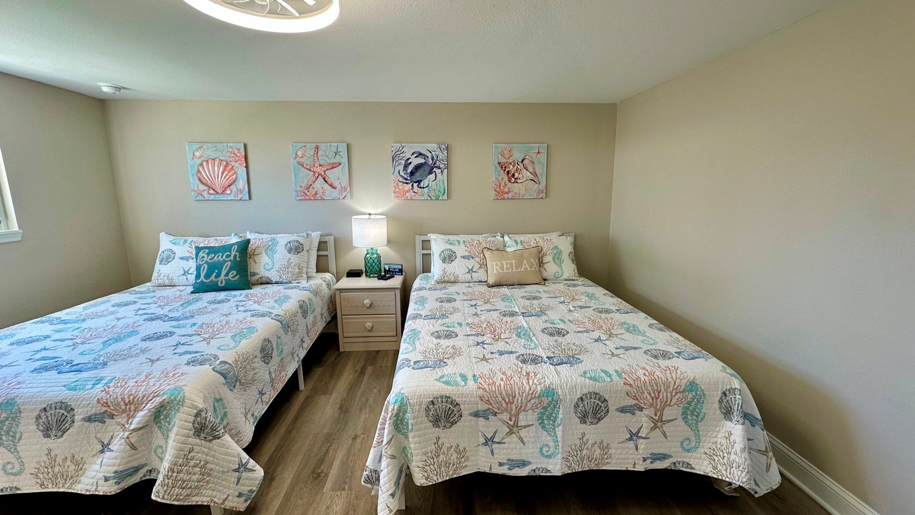 A bedroom with two beds, each with a sea-themed bedspread, a nightstand, a lamp, and four wall art pieces depicting sea life.