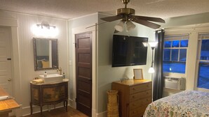 A bedroom with a wooden floor, a ceiling fan, a bed with a floral bedspread, a dresser, a mirror, and a television. - Jude's cozy & convenient downtown studio Apt. (High Point)