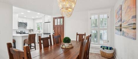 Un coin repas avec une table en bois, des chaises, un lustre et, au fond, une vue sur la cuisine.