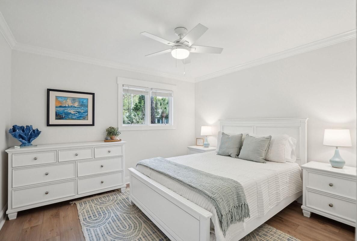 A bedroom with a bed, white nightstands, a ceiling fan, a window, and a framed picture on the wall.