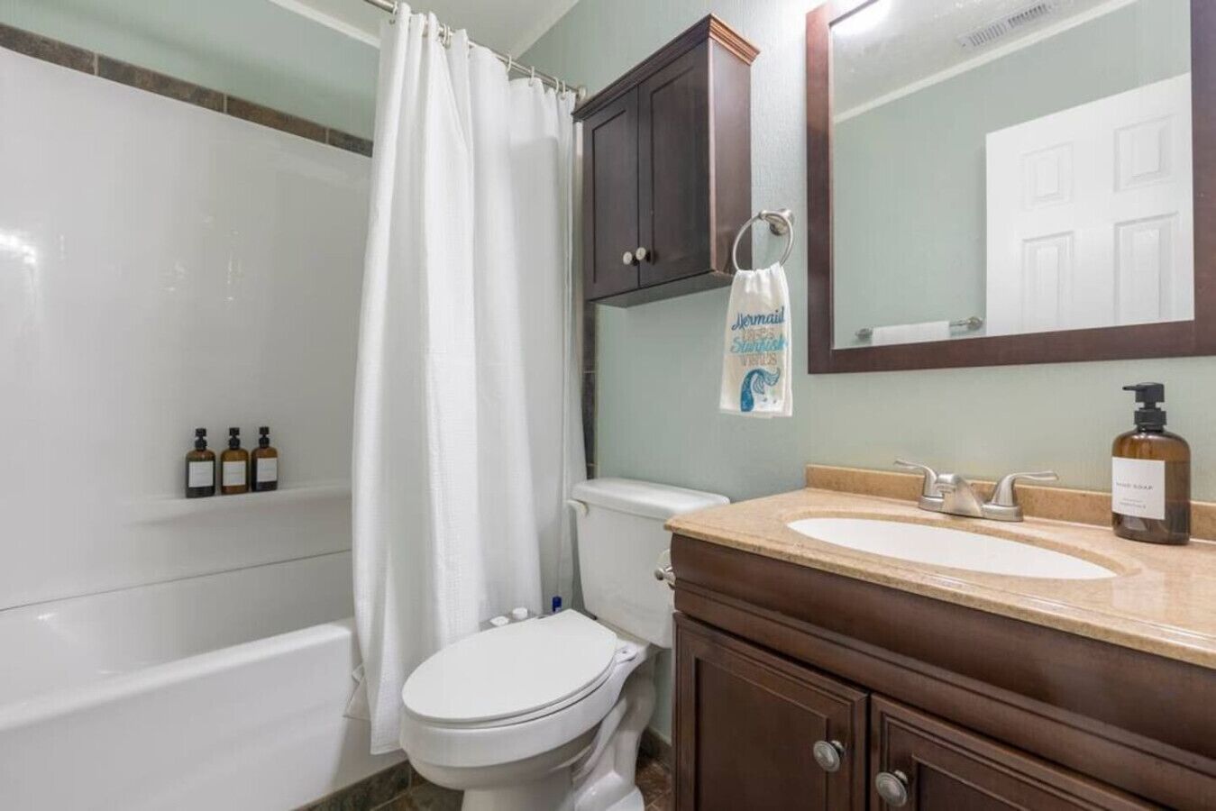 Une salle de bain équipée d’une baignoire, de toilettes et d’un meuble-lavabo avec un miroir. On y trouve des articles de toilette sur le meuble-lavabo, ainsi qu’un rideau de douche.
