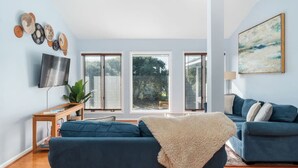 A modern living room with a blue sofa, a TV, and a large window with blinds. - Point Oasis|Golf Cart·The Point Beach·Sleeps 7·EI (Emerald Isle)