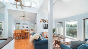 A spacious living area with a dining table, blue sofas, and large windows offering views of the outdoors. - Point Oasis|Golf Cart·The Point Beach·Sleeps 7·EI (Emerald Isle)