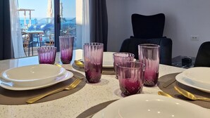 Ruokapöytä on katettu lautasilla, aterimilla ja laseilla, ja taustalla näkyy meri sekä tuoli.