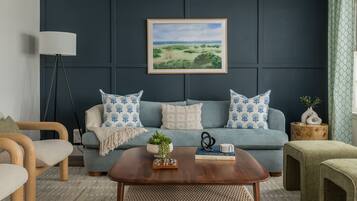 Un salon moderne avec un mur bleu foncé, un canapé bleu clair agrémenté de coussins à motifs, une table basse en bois et un tableau représentant un paysage, encadré.