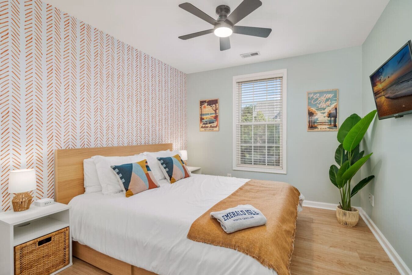 A bedroom with a bed, a ceiling fan, a window with blinds, a wall-mounted TV, and a potted plant.
