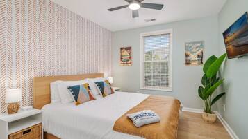 A bedroom with a bed, a ceiling fan, a window with blinds, a wall-mounted TV, and a potted plant.