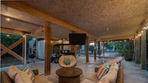 Un espace patio extérieur couvert, aménagé avec des meubles en osier, une table ronde et un téléviseur à écran plat fixé au plafond.