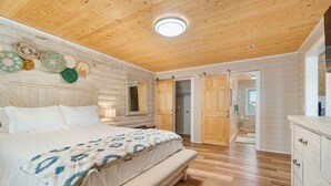 A bedroom with a wooden ceiling, a bed with a patterned blanket, a dresser, and a door leading to a bathroom. - Cabin by the Sea|1-Min Beach·Projector Room·Log Cabin EI (Emerald Isle)