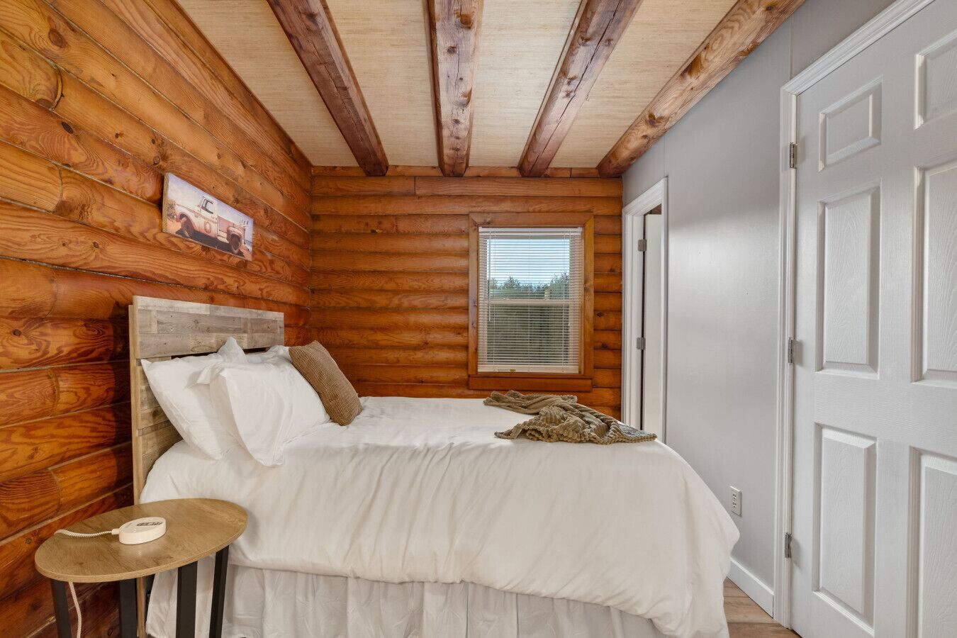 A bedroom with a wooden log wall, a bed with white bedding, a bedside table with a lamp, and a window with blinds.