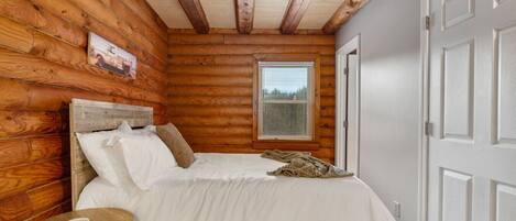 A bedroom with a wooden log wall, a bed with white bedding, a bedside table with a lamp, and a window with blinds.