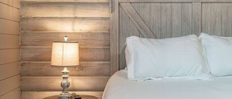 A bedroom with a wooden wall, a round table with a lamp, and a bed with white bedding.