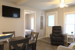 A modern living room with a flat-screen TV, a dark armchair, a dining table with chairs, and a small side table. - Spacious 3BR Retreat | Perfect for Families! (Indian River)