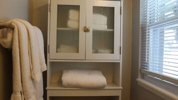 Une salle de bain avec un meuble blanc contenant des serviettes, un porte-serviettes et une fenêtre avec des stores.