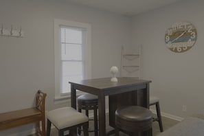 Quarto com mesa de madeira, duas cadeiras, um banco, um relógio e uma janela com persianas.