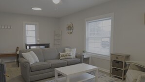 A living room with a grey sofa, a white coffee table, a small side table, a window with blinds, and a wall clock. - Cozy Riverfront Getaway | 1BR + Sleeper Sofa (Indian River)
