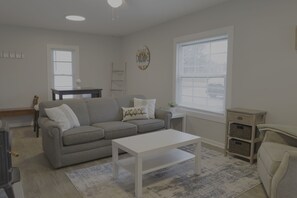 A living room with a grey sofa, a white coffee table, a small side table, a window with blinds, and a wall clock.