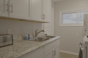 A kitchen with white cabinets, a granite countertop, a stainless steel sink, and a window with a view of trees. - Cozy Riverfront Getaway | 1BR + Sleeper Sofa (Indian River)