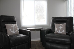 Deux fauteuils en cuir noir, ornés de coussins à motifs d’ancre, sont placés devant une fenêtre munie de stores.