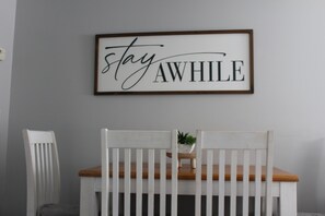 Un cartel en marco con la frase 'stay awhile' en una pared, dos sillas blancas con asientos de madera y una peque ña planta en maceta sobre una mesa de madera.
