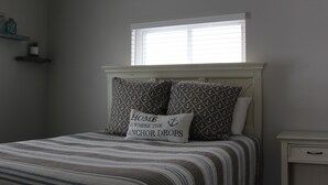 Een slaapkamer met een bed, kussens, een nachtkastje, een raam en planken met decoratieve objecten.
