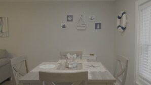 A dining area with a table set for two, a white chair, a small white table, and a window with blinds. - 1BR Hideaway | Steps from Indian River (Indian River)