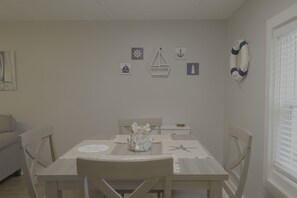 A dining area with a table set for two, a white chair, a small white table, and a window with blinds. - 1BR Hideaway | Steps from Indian River (Indian River)