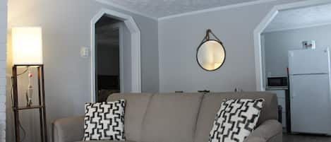 A living room with a beige sofa, patterned throw pillows, a wall-mounted mirror, and a side table with a lamp.