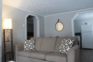 A living room with a beige sofa, patterned throw pillows, a wall-mounted mirror, and a side table with a lamp.