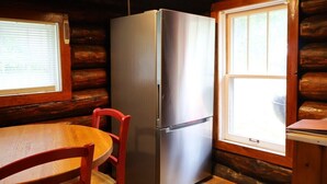 A compact kitchen with a stainless steel refrigerator, wooden walls, a wooden dining table with a red chair, and a window providing natural light. - Rustic Riverfront Getaway (Indian River)