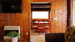 A wooden cabin interior with a TV mounted on the wall, a fireplace, a dining area with red chairs, and a window providing natural light. - Rustic Riverfront Getaway (Indian River)