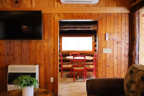 L’intérieur d’un chalet en bois est doté d’un téléviseur fixé au mur, d’une cheminée, d’un coin repas avec des chaises rouges et d’une fenêtre qui laisse entrer la lumière naturelle.