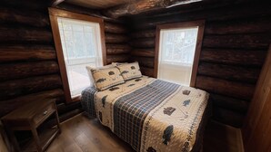 A cozy cabin bedroom with a wooden floor, a bed with a quilted cover, two windows with blinds, and a ceiling light. - Rustic Riverfront Getaway (Indian River)