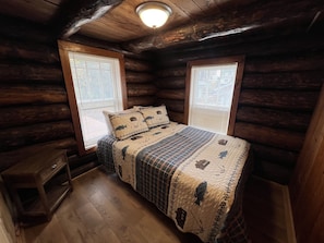 A cozy cabin bedroom with a wooden floor, a bed with a quilted cover, two windows with blinds, and a ceiling light.