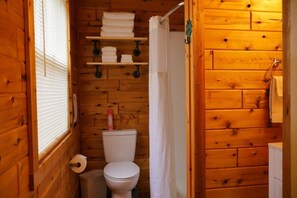 A wooden cabin bathroom with a toilet, shower curtain, and shelves holding towels. - Riverside Escape | Perfect for Families & Friends (Indian River)