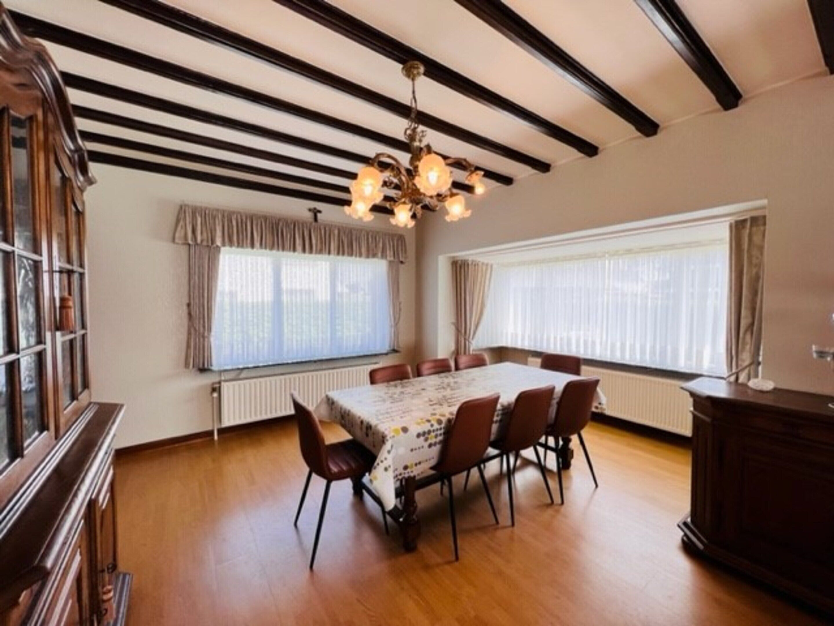 A dining room with a wooden table, six chairs, a chandelier, and large windows with curtains.