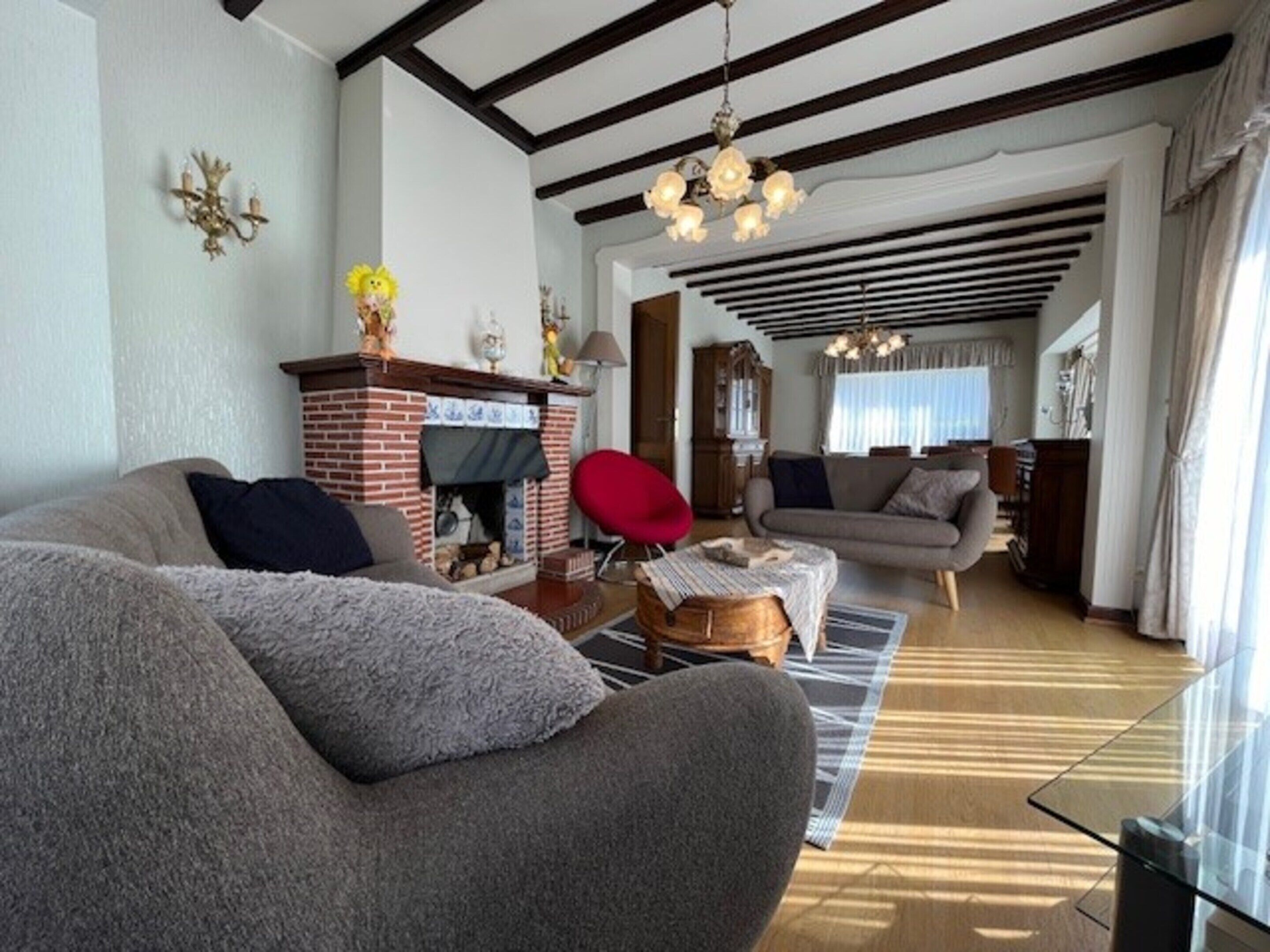 A living room with a fireplace, a red chair, and a sofa. There is a dining area with a table and chairs in the background.