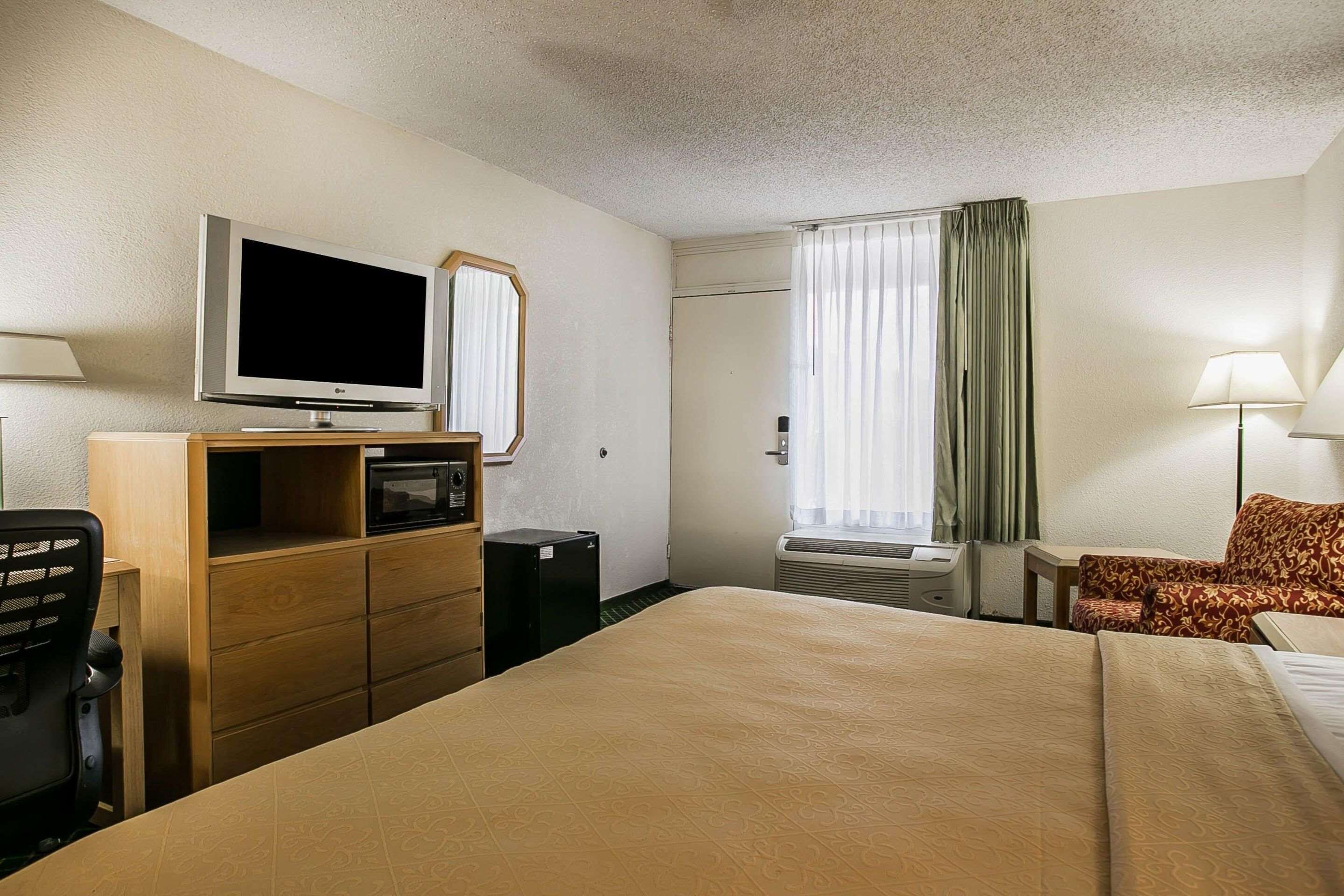 Habitación de hotel con cama, escritorio con computadora, televisor, espejo, ventana con cortinas y lámpara.
