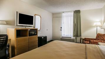 A hotel room with a bed, a desk with a computer, a television, a mirror, a window with curtains, and a lamp.