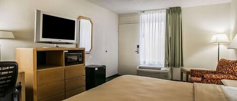 Habitación de hotel con cama, escritorio con computadora, televisor, espejo, ventana con cortinas y lámpara.
