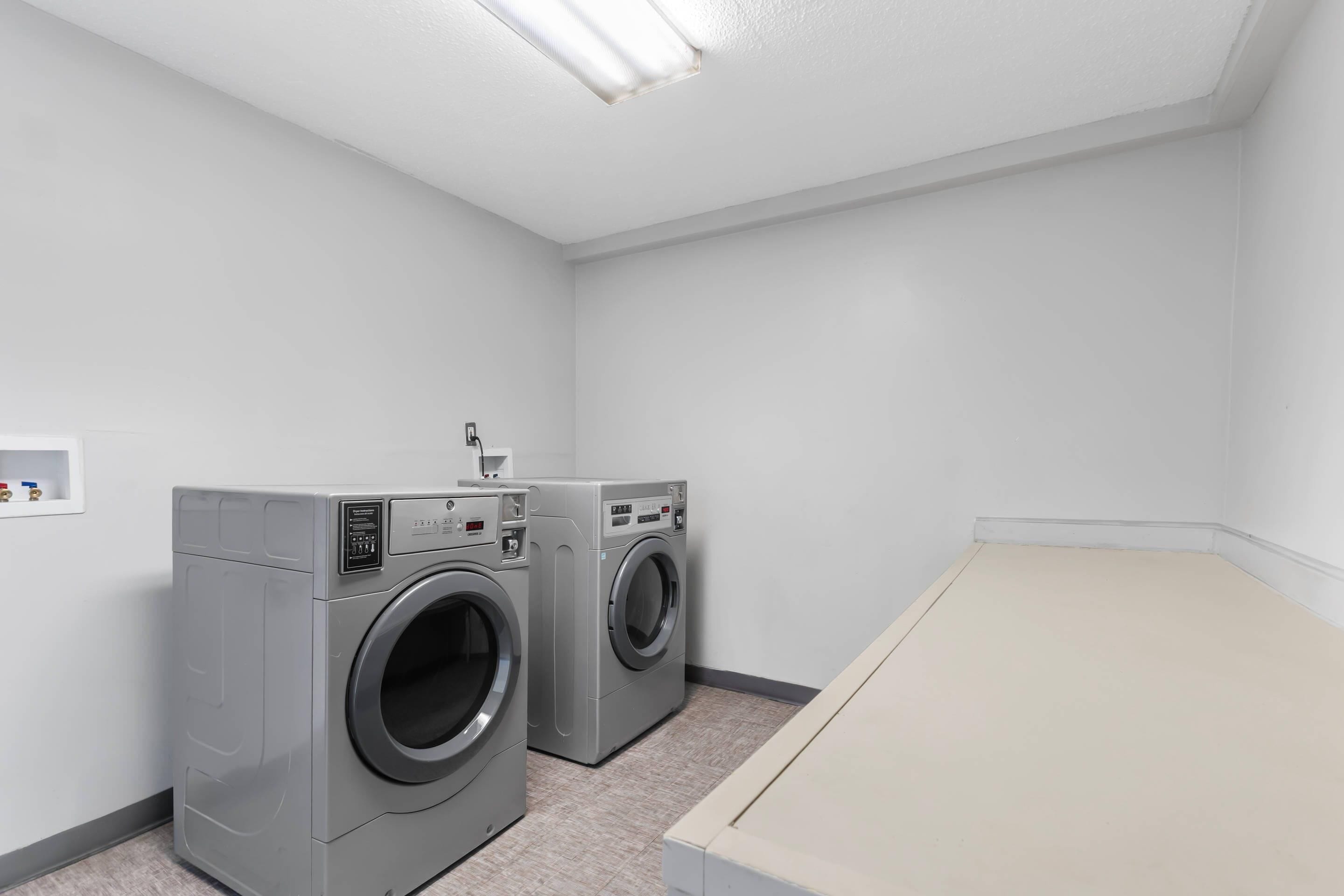 A laundry room with two front-loading washing machines, a small shelf with items, and a bench.