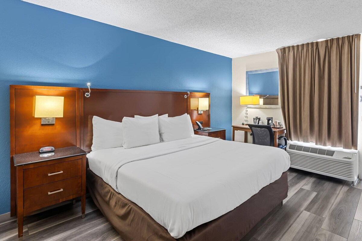 Quarto de hotel com uma cama grande, uma escrivaninha com cadeira, uma luminária de mesa e uma janela com cortinas.