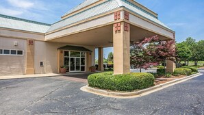 A hotel with a covered entrance, a parking lot, and landscaped areas with shrubs and a tree.