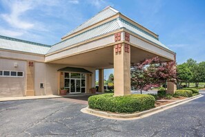 A hotel with a covered entrance, a parking lot, and landscaped areas with shrubs and a tree.