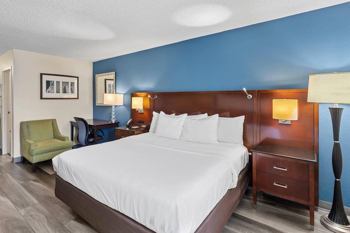 Quarto de hotel com uma cama grande, uma escrivaninha com cadeira, uma poltrona verde, duas mesas de cabeceira com luminárias e um espelho na parede.