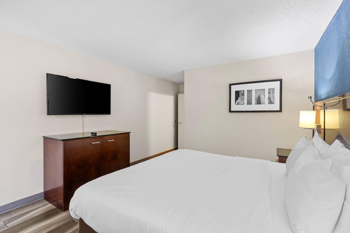 Quarto de hotel com cama, televisão de tela plana, penteadeira de madeira, fotografia emoldurada na parede e abajur.