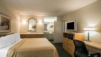A hotel room with a bed, a desk with a computer and a TV, a chair, a microwave, a refrigerator, and a bathroom visible through an open door.