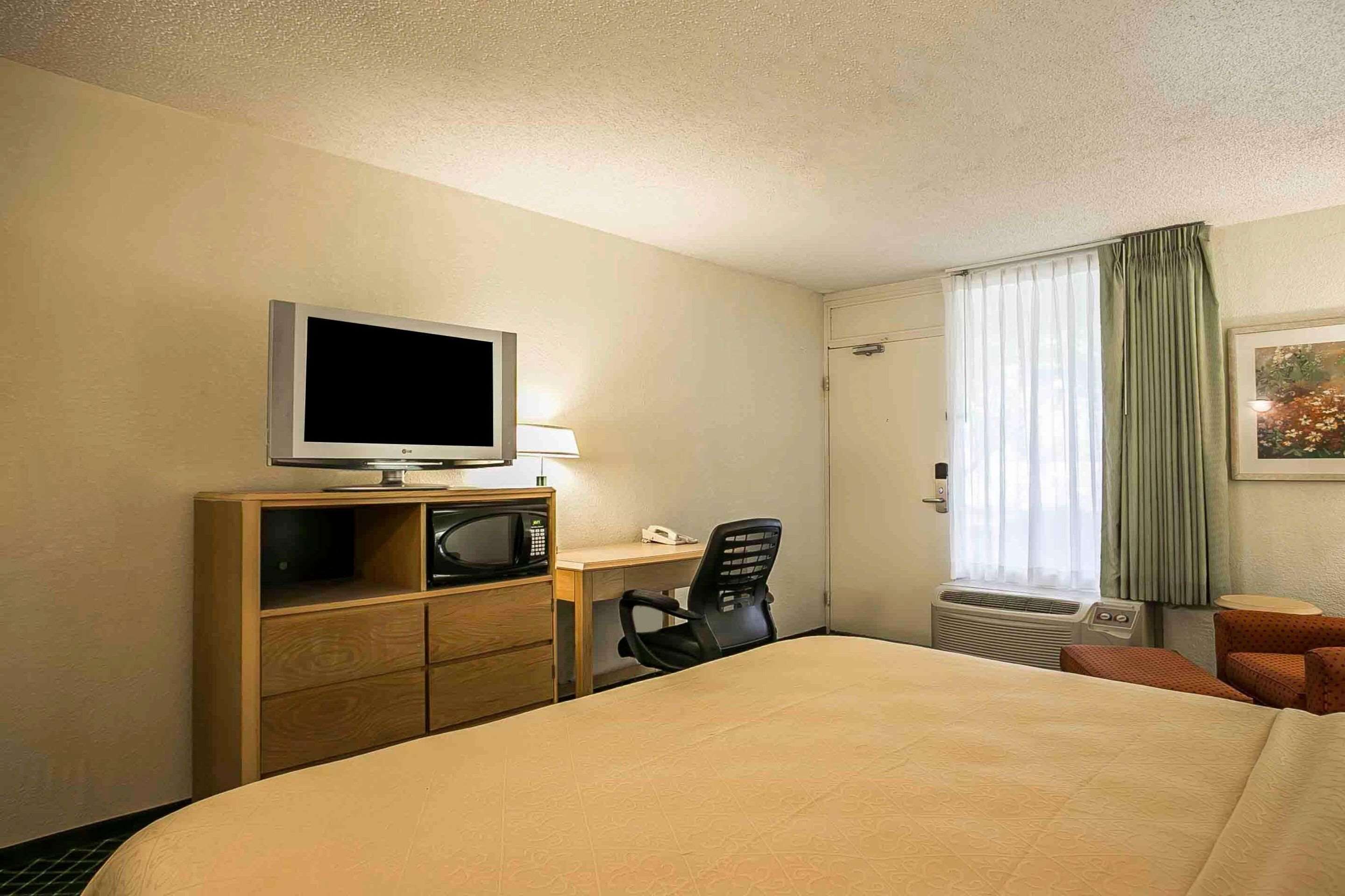 Een hotelkamer met een bed, een bureau met een computer, een televisie, een magnetron, een stoel en een raam met gordijnen.