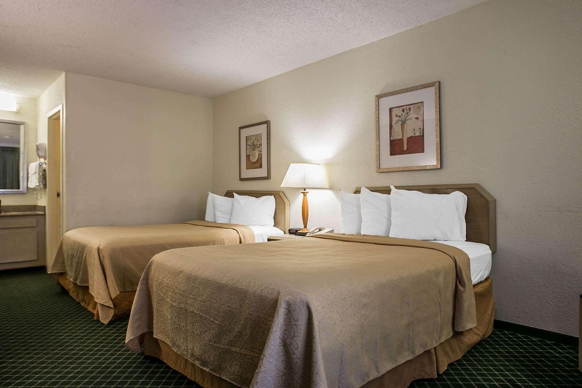 Quarto de hotel com duas camas, uma mesa de cabeceira com abajur e um quadro na parede.